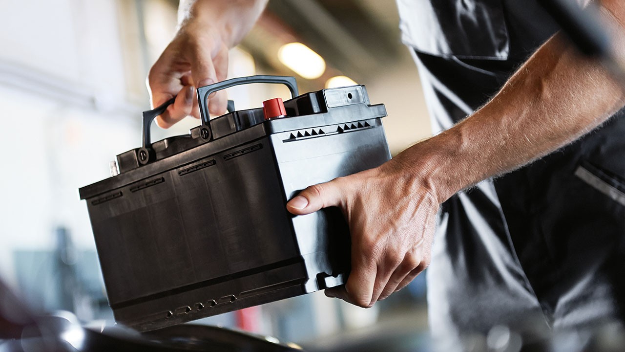 Mechanic installing a car battery under the hood in an auto repair shop. Close-up of a technician’s hands holding a black automotive battery with visible terminals, wearing a MINI Service uniform.
